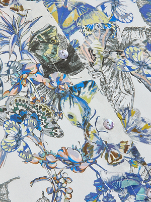 A close-up of white fabric with colorful butterfly and floral prints in blue, green, and yellow shades, featuring two white buttons—a detail from the elegant Tyler Silk Blouse.