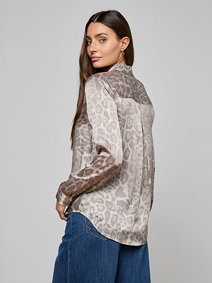 A woman with long brown hair wears the L'AGENCE Tyler Silk Blouse in light gray leopard print and blue jeans, standing sideways and looking over her shoulder against a plain gray background.