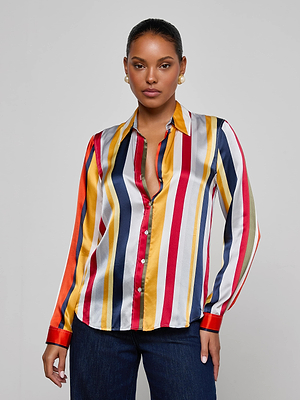 A woman wears the L'AGENCE Tyler Silk Blouse featuring red, yellow, white, blue, and gray vertical stripes, paired with dark jeans. She stands against a light gray background with pearl earrings and a confident expression.