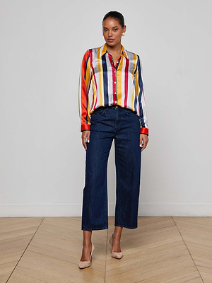 A woman stands indoors against a white wall, wearing the L’AGENCE Tyler Silk Blouse with colorful stripes, paired with dark blue wide-leg jeans and beige heels on a light wood floor.