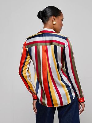 A woman stands with her back to the camera, wearing the L'AGENCE Tyler Silk Blouse featuring bold multi-colored vertical stripes. Her hair is in a bun, and she pairs it with gold circular earrings and dark pants.