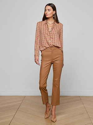 A woman poses indoors against a plain light wall, wearing the L'AGENCE Holly Blouse with a horse bit print, paired with tan high-waisted pants and heels. She has long brown hair, jewelry, and one hand in her pocket.