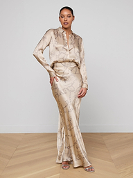 A woman stands against a plain white background, wearing the L’AGENCE Tyler Silk Blouse in a light beige chain print with a matching floor-length skirt and open-toe sandals. She has one hand on her hip and looks confidently at the camera.