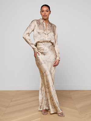 A woman stands on a wooden floor, confidently posing in strappy sandals and a cream-colored Tyler Silk Blouse from L'AGENCE, paired with a matching subtly patterned long skirt.