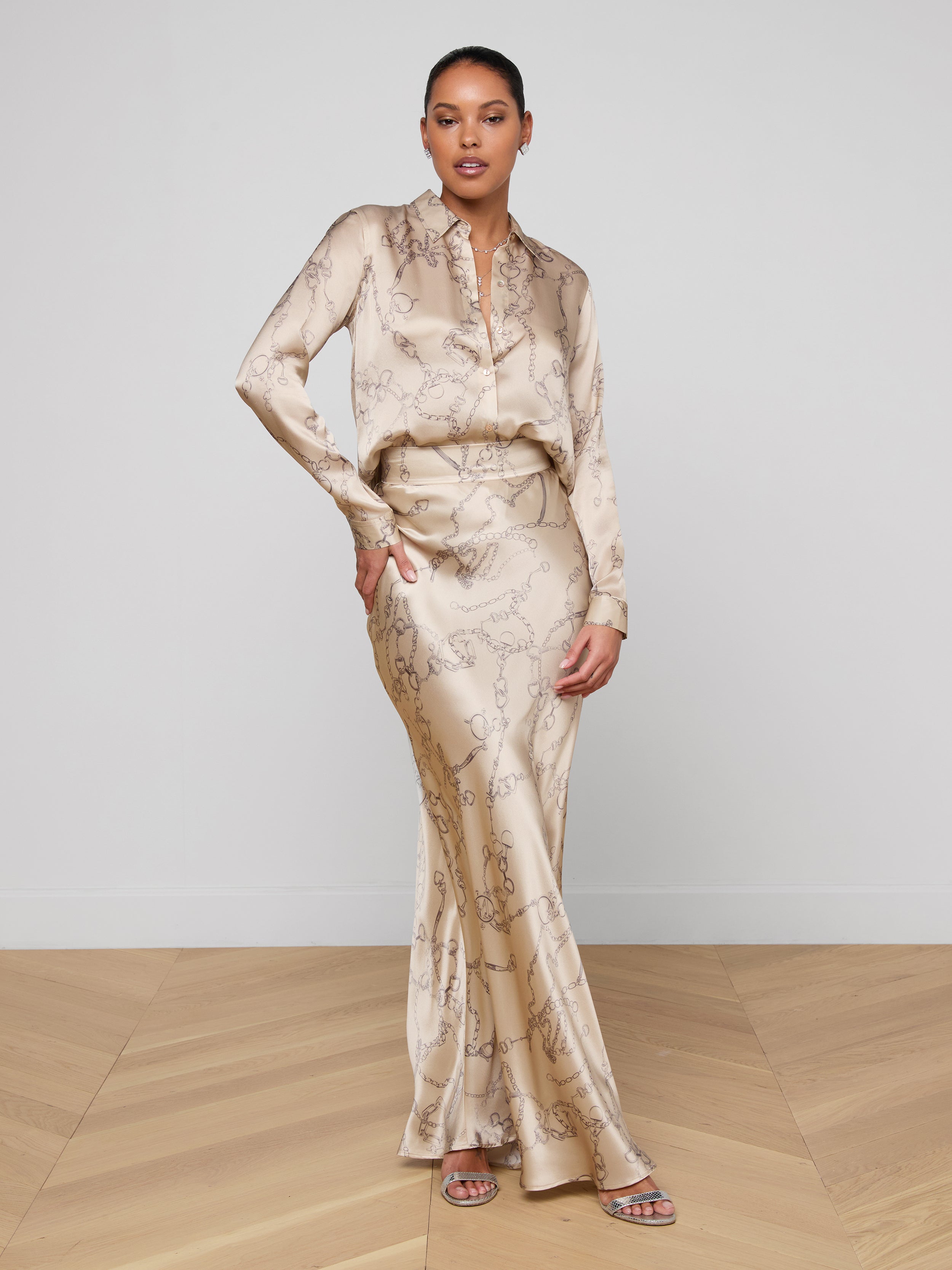 A woman stands against a plain white background, wearing the L’AGENCE Tyler Silk Blouse in a light beige chain print with a matching floor-length skirt and open-toe sandals. She has one hand on her hip and looks confidently at the camera.