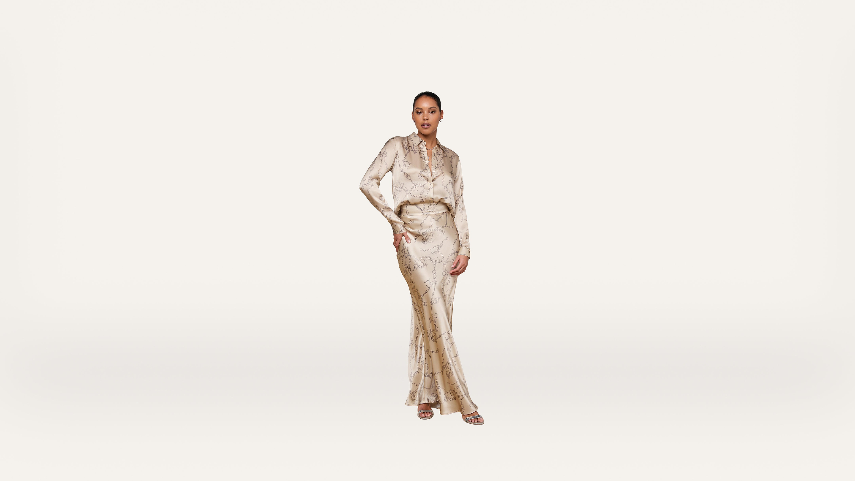 Woman stands confidently wearing a light-colored, full-length, patterned dress with long sleeves and a belted waist, set against a plain white background.