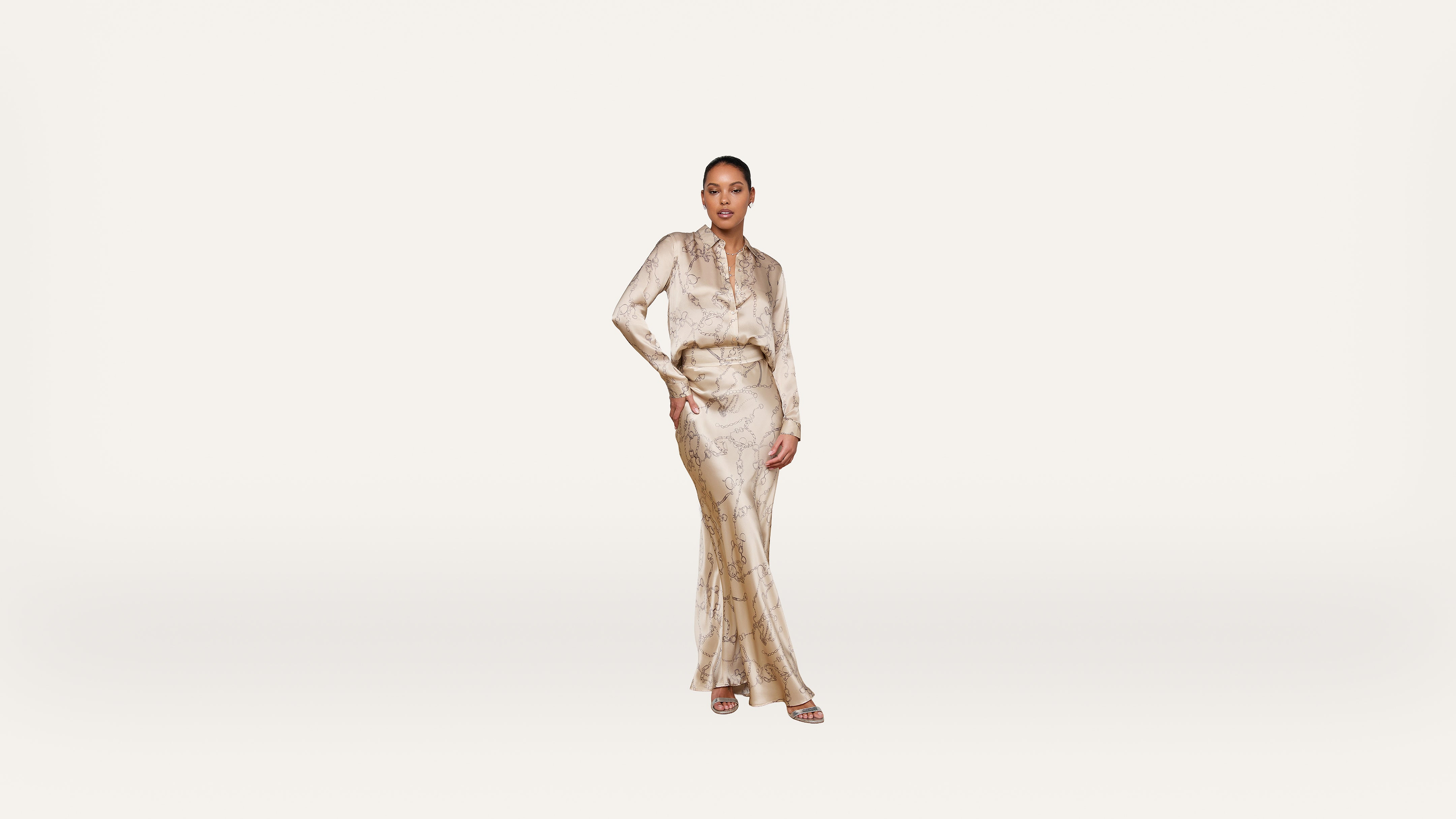 Woman stands confidently wearing a light-colored, full-length, patterned dress with long sleeves and a belted waist, set against a plain white background.
