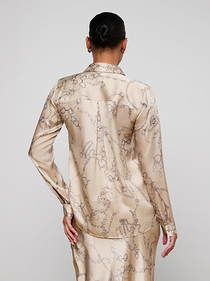 A woman with dark hair in a bun is shown from the back, wearing the shiny chain print L'AGENCE Tyler Silk Blouse with a matching skirt. She rests one hand on her hip against a plain white background.