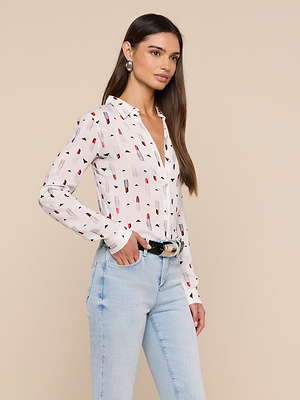 A woman with long brown hair wears the Holly Blouse, a slim fit, long sleeve button down with a lipstick print, paired with light blue high-waisted jeans, a black belt, and large silver earrings against a beige background.