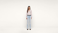 A woman with long brown hair stands facing forward against a plain white background, wearing a white blouse with a heart pattern, light blue jeans, a black belt, and black shoes.