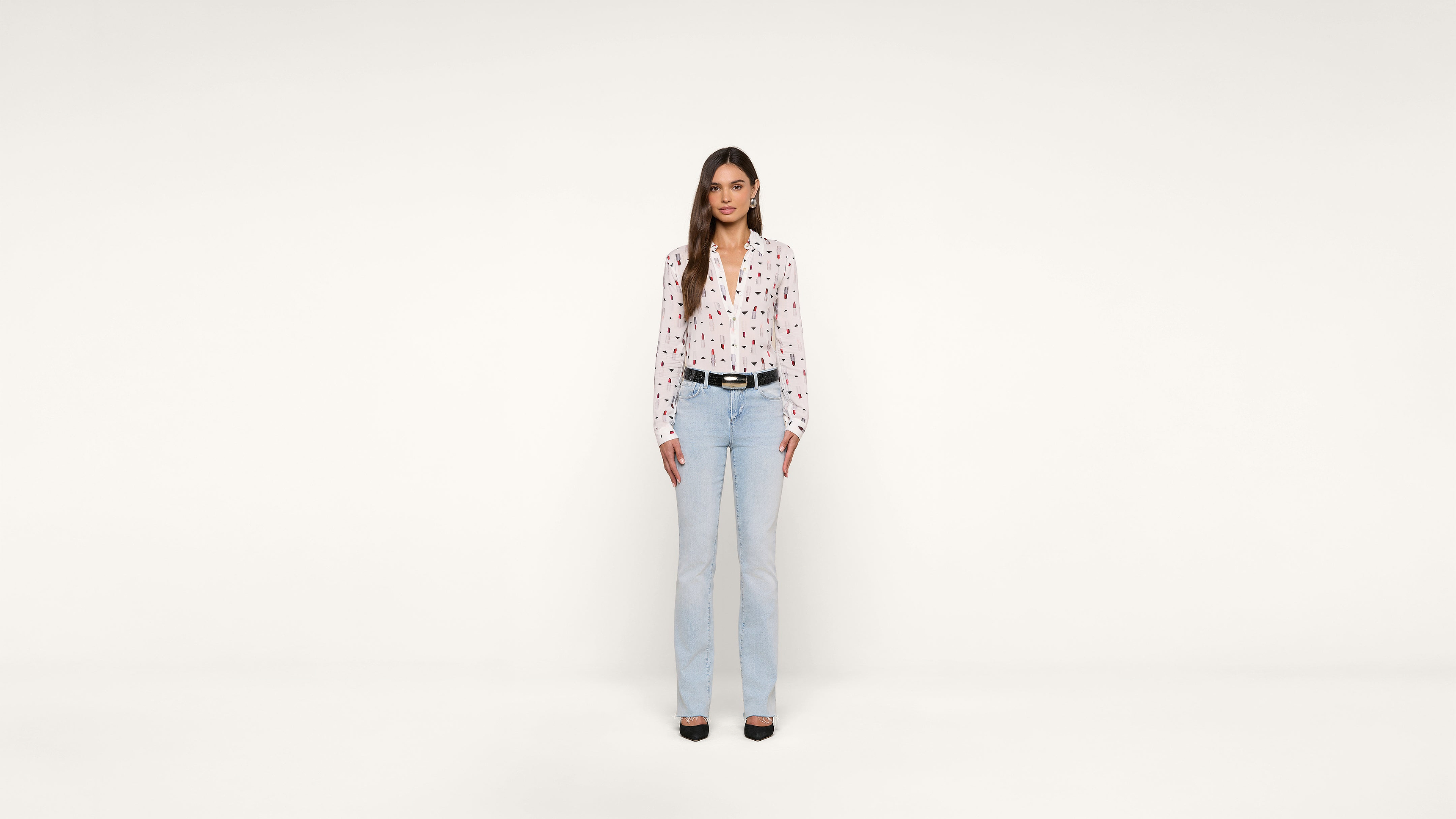 A woman with long brown hair stands facing forward against a plain white background, wearing a white blouse with a heart pattern, light blue jeans, a black belt, and black shoes.