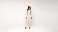 A woman stands against a plain white background, wearing sunglasses, a patterned blouse, beige trousers, a brown belt, and high-heeled sandals.