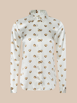 The Tyler Silk Blouse is a white, long-sleeve, collared button-down with brown and gold heart prints evenly spaced across the fabric, shown on a beige background.
