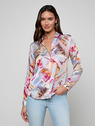 A woman stands and smiles against a light background, wearing the Tyler Silk Blouse with a vibrant pink postcard print and light blue jeans.