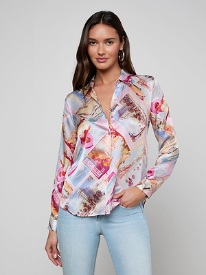 A woman with long brown hair wears the LAGENCE Tyler Silk Blouse in a colorful pink postcard print and light blue jeans, posing against a plain light gray background with a slight smile, one hand by her side.