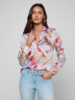 A woman with long brown hair wears the L’AGENCE Tyler Silk Blouse in a pink postcard print and light blue jeans, standing against a plain gray background and looking confidently at the camera with one hand touching her collar.