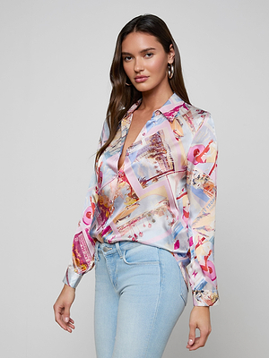 A woman poses confidently against a plain light gray background, wearing the LAGENCE Tyler Silk Blouse with a vibrant pink postcard print and light blue jeans.
