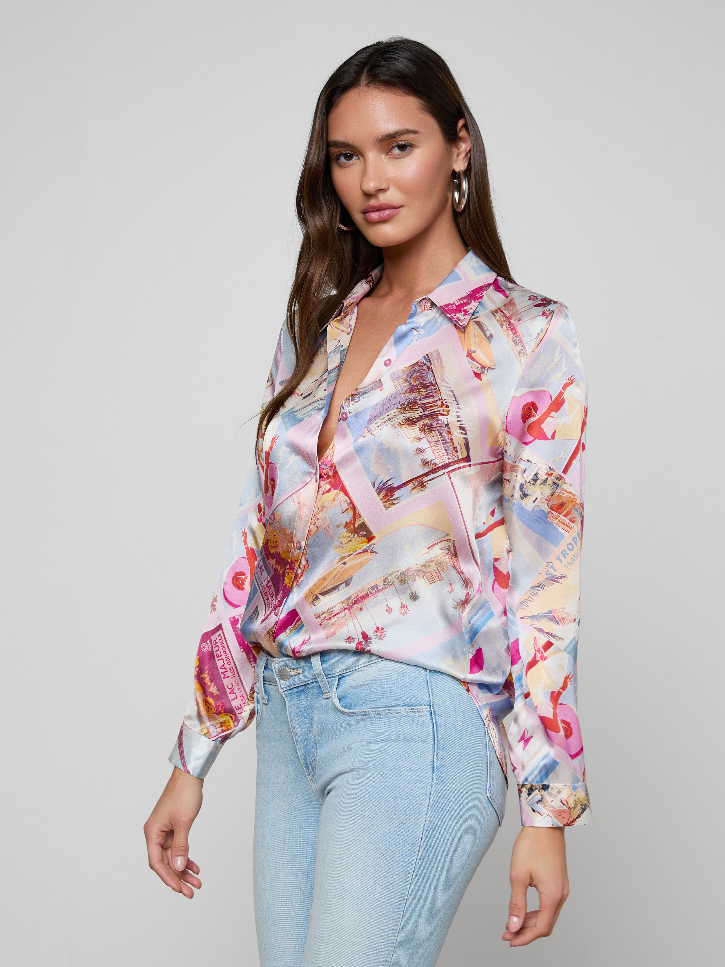 A woman stands against a plain, light background, looking at the camera with a slight smile. She is wearing the Tyler Silk Blouse—a colorful, pink postcard print long-sleeve silk button-down—paired with light blue jeans.