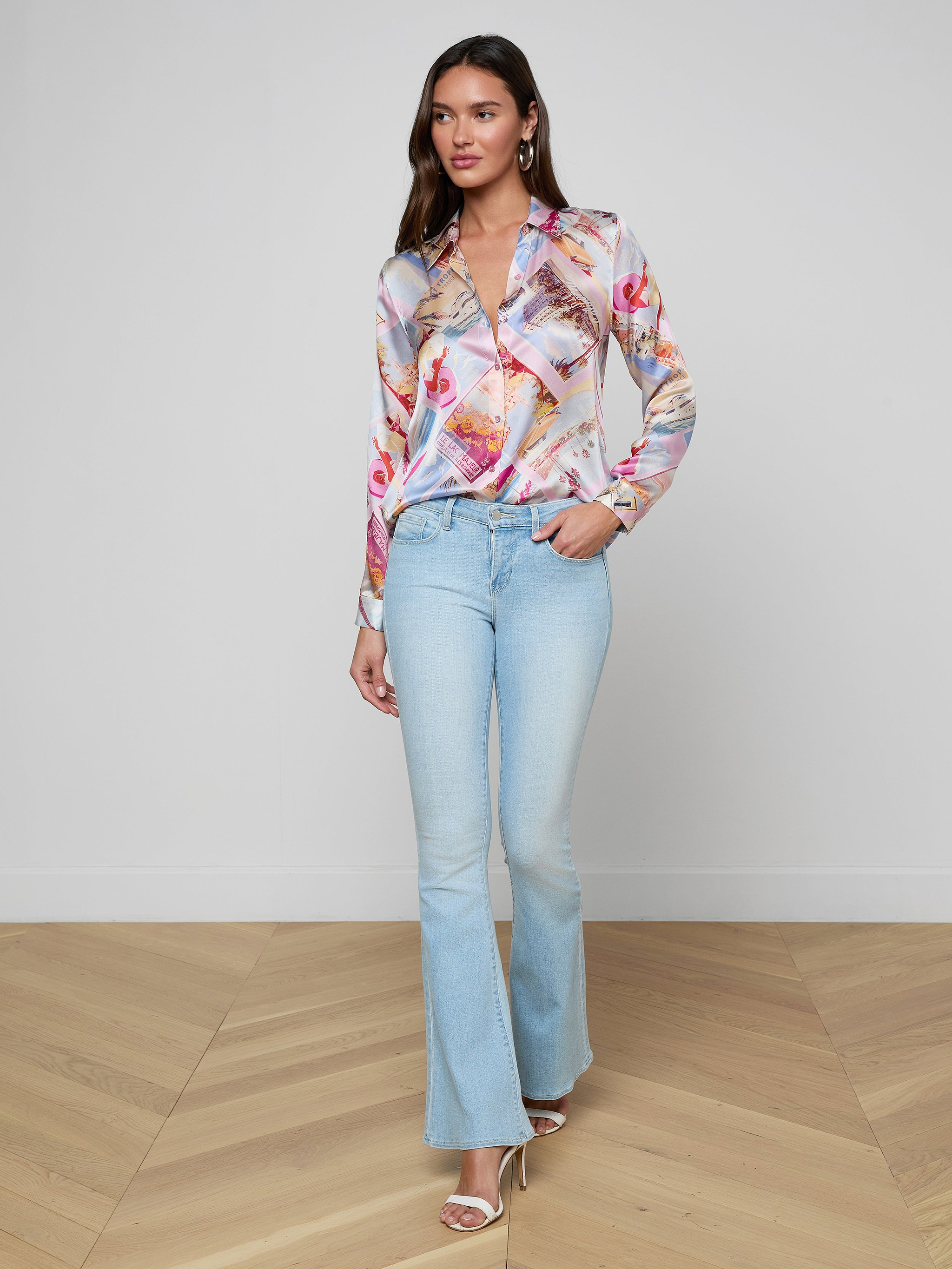 A woman stands confidently against a plain white background, wearing the colorful Tyler Silk Blouse from L’AGENCE paired with light blue flared jeans, her hand in her pocket and open-toed heels.