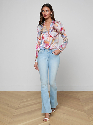 A woman with long brown hair stands indoors against a plain background wearing the Tyler Silk Blouse from LAGENCE in a pink postcard print, paired with light blue flared jeans and heeled sandals, posing with one hand in her pocket.