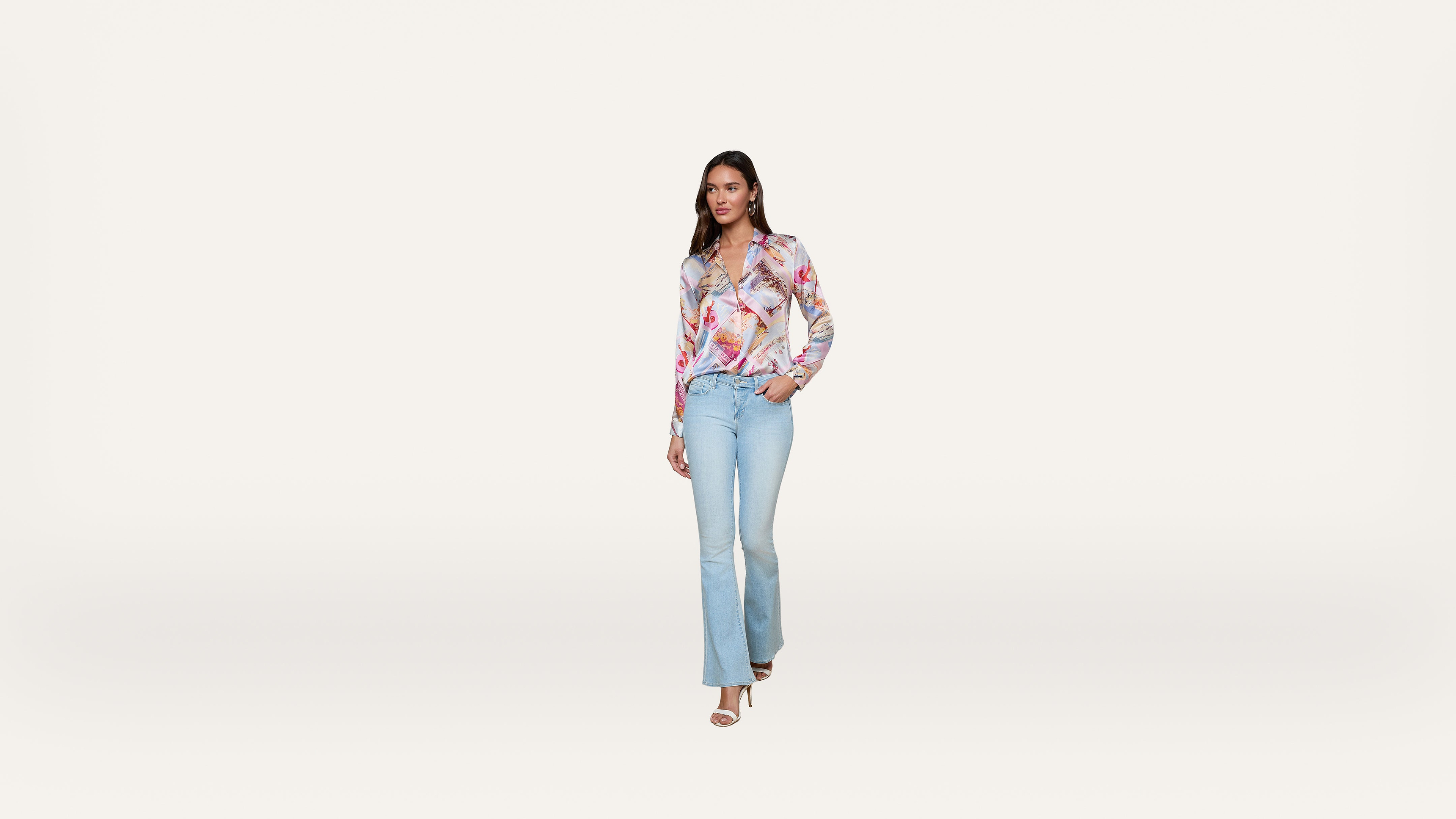 A woman in a colorful blouse and light blue jeans stands against a plain white background, with one hand in her pocket and the other relaxed by her side.