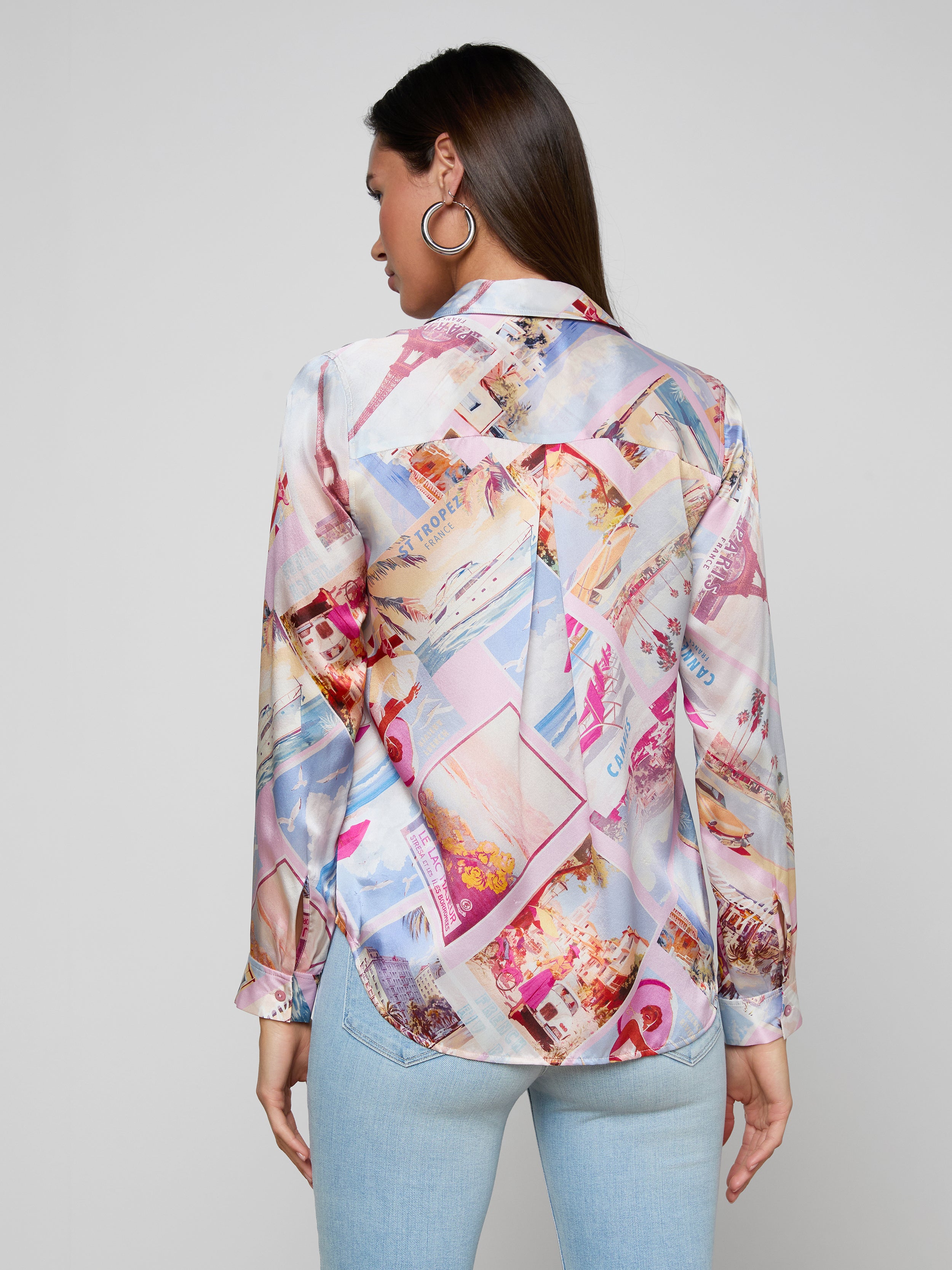 A woman faces away, wearing the Tyler Silk Blouse by L’AGENCE featuring a pink postcard print, paired with light blue jeans. Her dark hair is styled down, and she accessorizes with large hoop earrings. The background is plain and light.