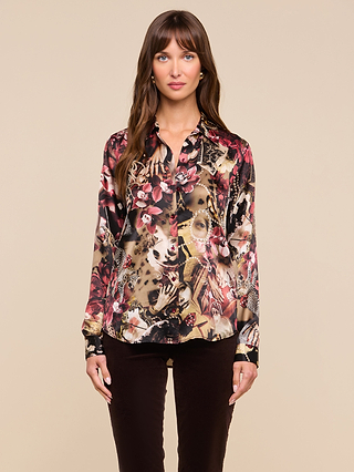 A woman stands facing forward, wearing the Tyler Silk Blouse—a long-sleeve button-down top featuring a colorful statement print of floral and animal motifs—paired with dark pants, against a plain beige background.