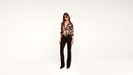 Woman wearing sunglasses, a colorful floral blouse, and dark flared pants stands against a plain white background.