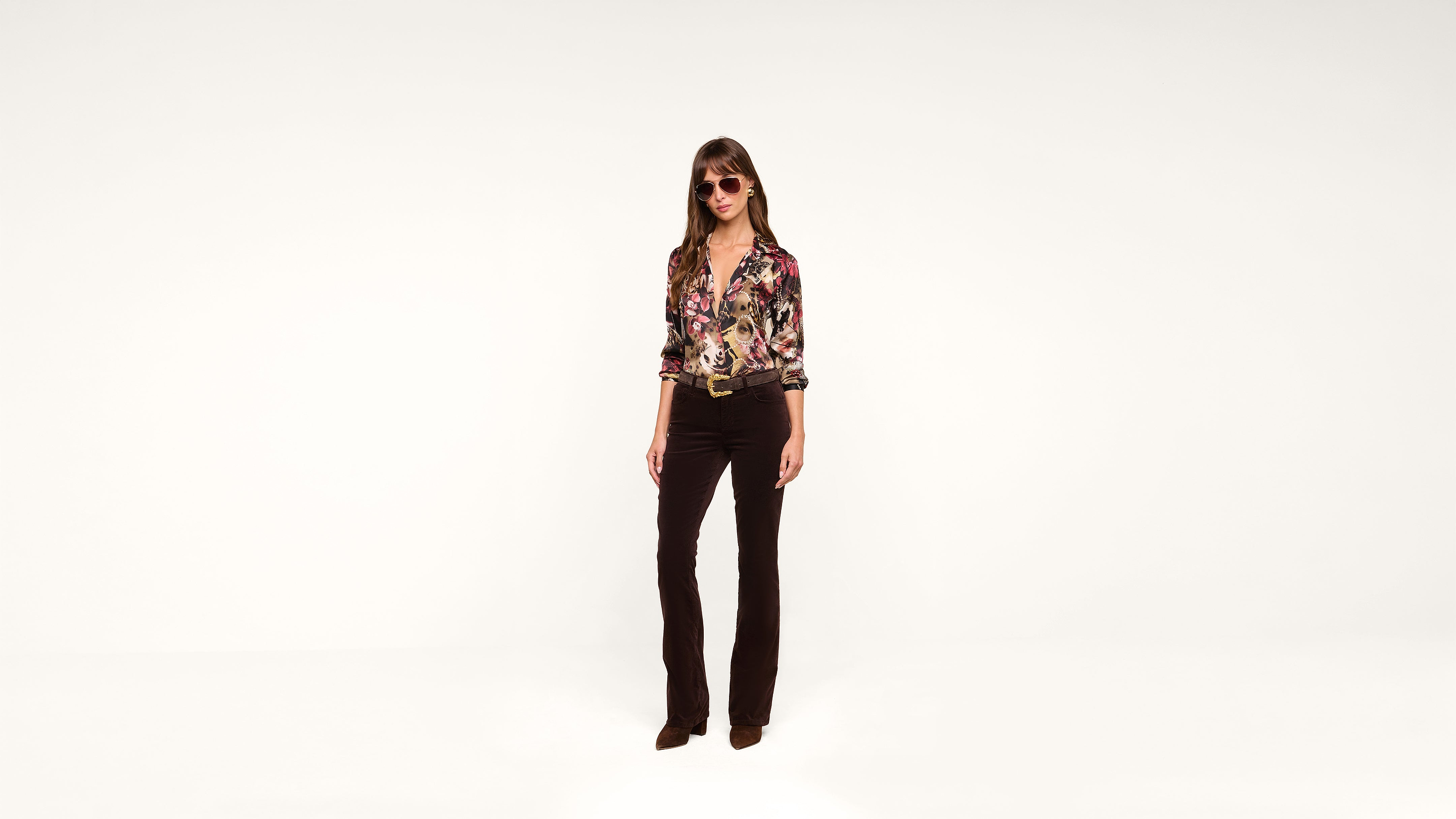 Woman wearing sunglasses, a colorful floral blouse, and dark flared pants stands against a plain white background.