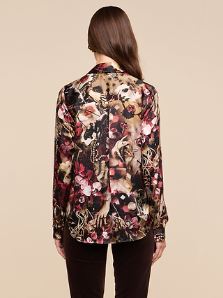 A person with long brown hair, shown from the back, wears the Tyler Silk Blouse—a statement long-sleeve shirt adorned with an elaborate floral and animal pattern in shades of red, pink, brown, and beige—paired with dark pants against a beige background.