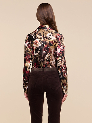 A woman with long brown hair stands with her back to the camera against a beige background, wearing the Tyler Silk Blouse—a long-sleeve, floral-patterned top—paired with dark brown pants.