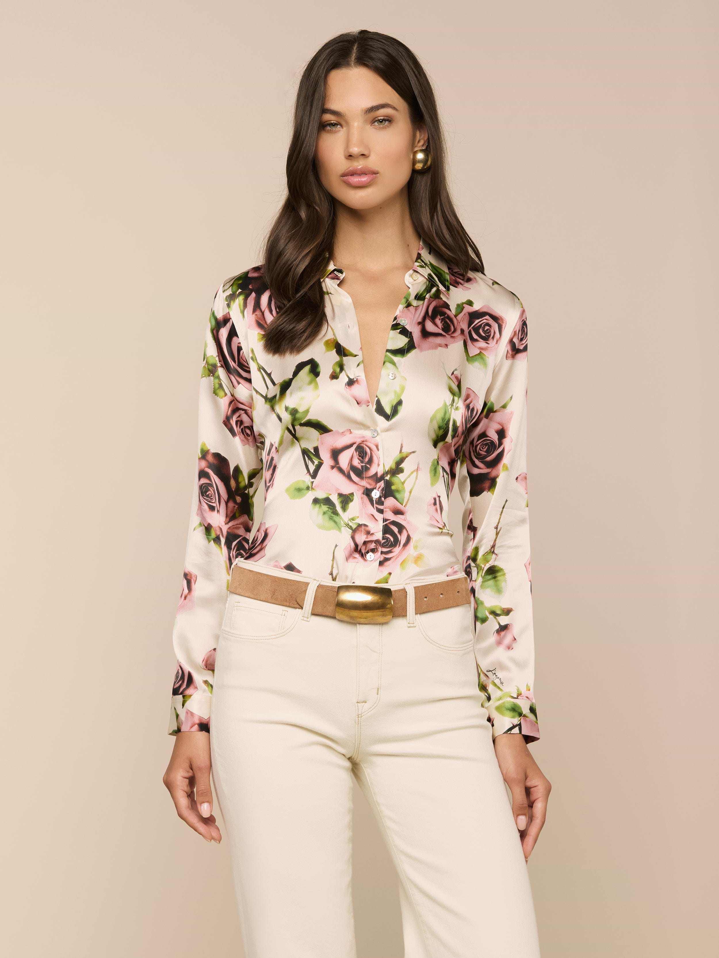 Una donna dai lunghi capelli castani indossa la blusa di seta LAGENCE Tyler con stampa di rose color malva e pantaloni color crema, su uno sfondo beige semplice e con unespressione neutra.