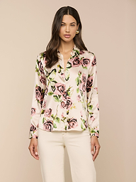 Una donna dai lunghi capelli scuri indossa la blusa di seta Tyler con stampa di rose color malva, abbinata a pantaloni bianchi, su un semplice sfondo beige.