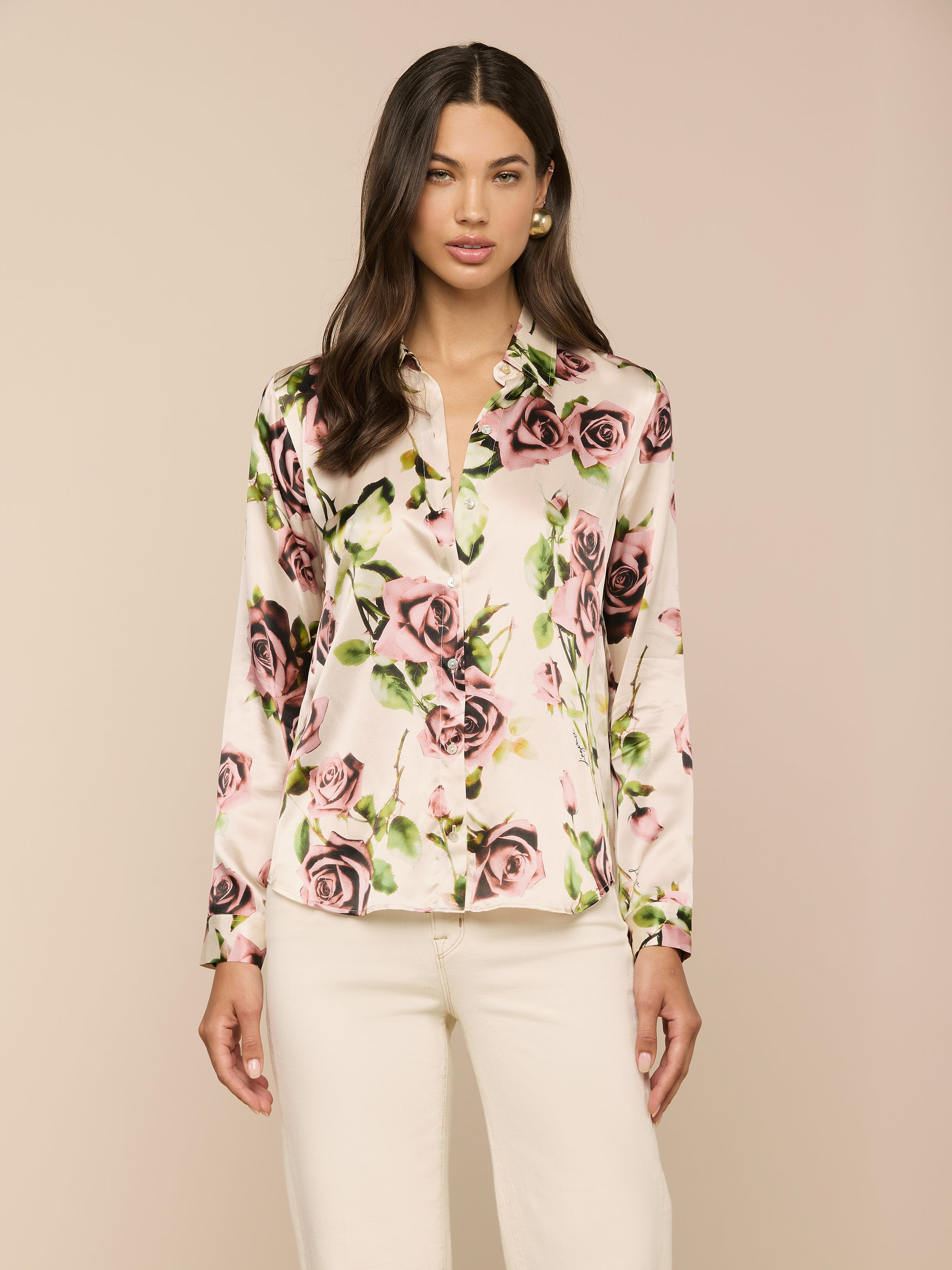 Una donna dai lunghi capelli scuri indossa la blusa di seta Tyler con stampa di rose color malva, abbinata a pantaloni bianchi, su un semplice sfondo beige.