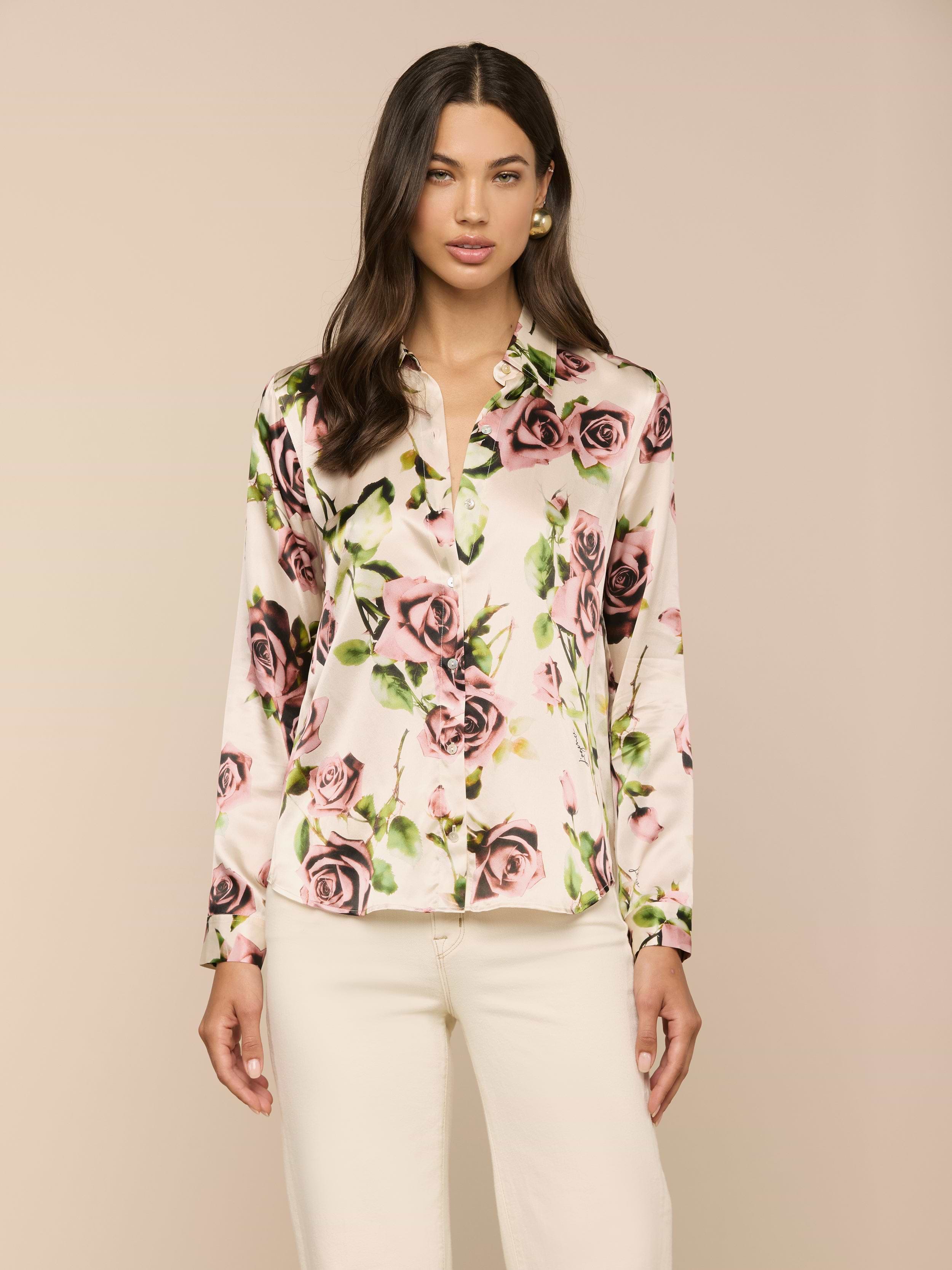 Una donna dai lunghi capelli scuri indossa la blusa di seta Tyler con stampa di rose color malva, abbinata a pantaloni bianchi, su un semplice sfondo beige.