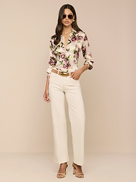 Una donna che indossa occhiali da sole, la blusa di seta Tyler con stampa rosa malva, pantaloni a vita alta color crema e sandali con tacco beige si trova su uno sfondo beige con una mano in tasca.