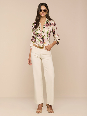 A woman wearing sunglasses, the Tyler Silk Blouse in mauve rose print, cream high-waisted pants, and beige heeled sandals stands against a beige background with one hand in her pocket.