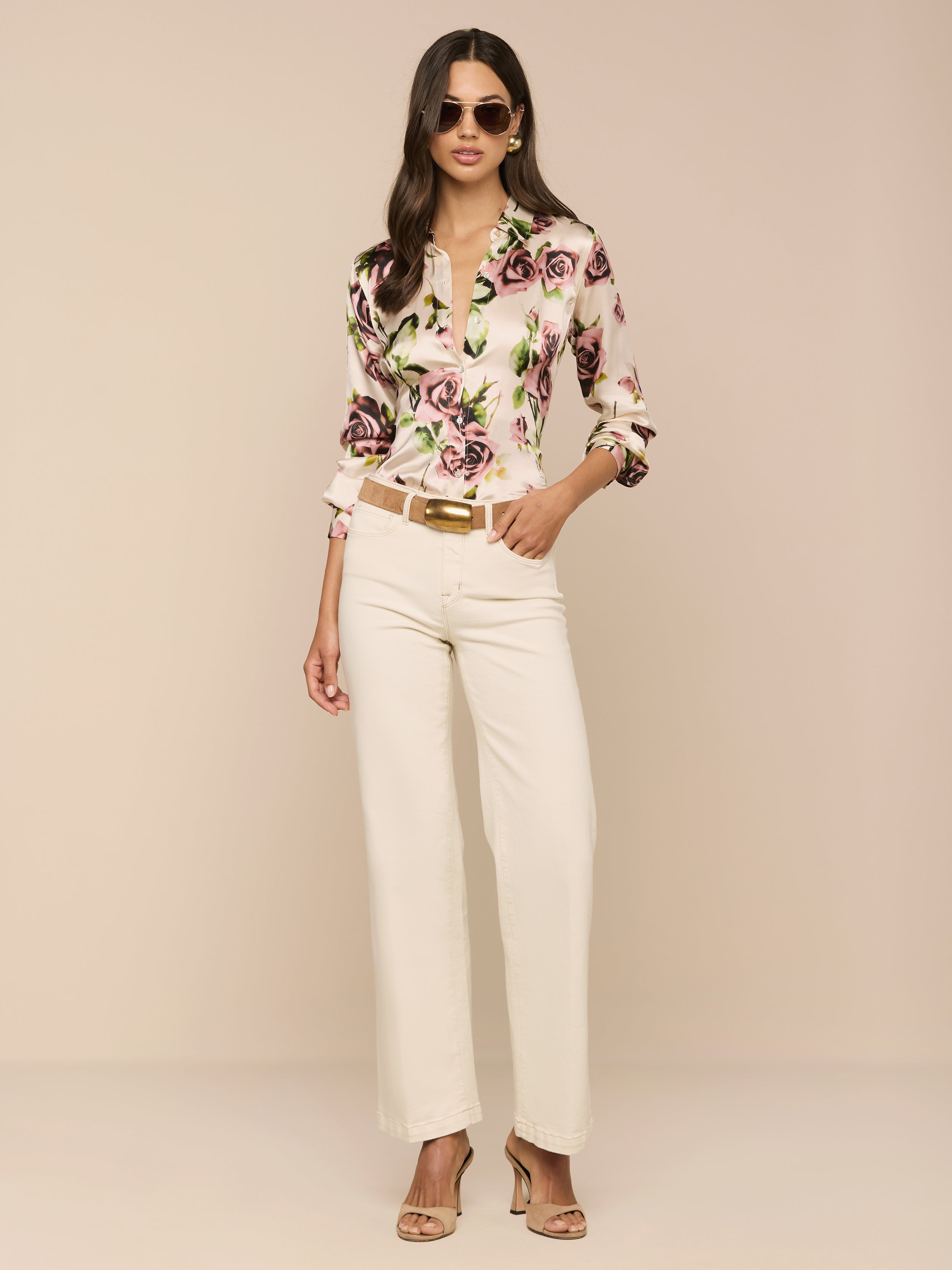 Una donna che indossa occhiali da sole, la blusa di seta Tyler con stampa rosa malva, pantaloni a vita alta color crema e sandali con tacco beige si trova su uno sfondo beige con una mano in tasca.
