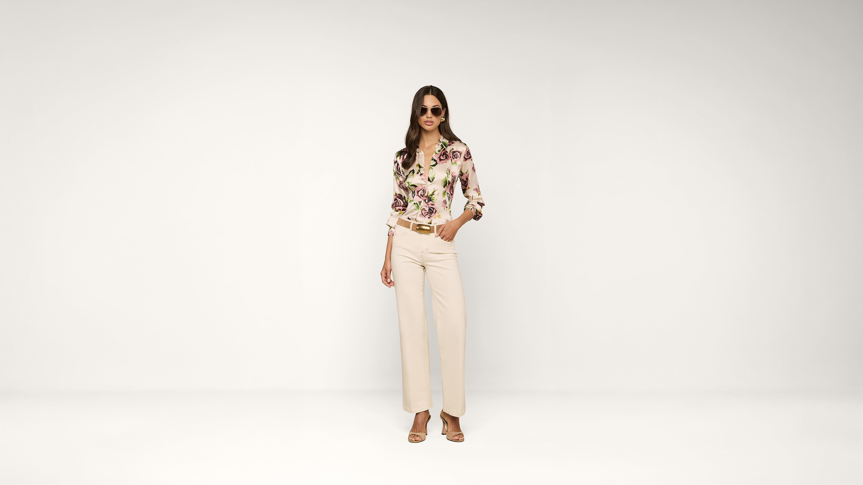 A woman wearing sunglasses, a floral blouse, cream wide-leg pants, and heeled sandals stands confidently against a plain white background.