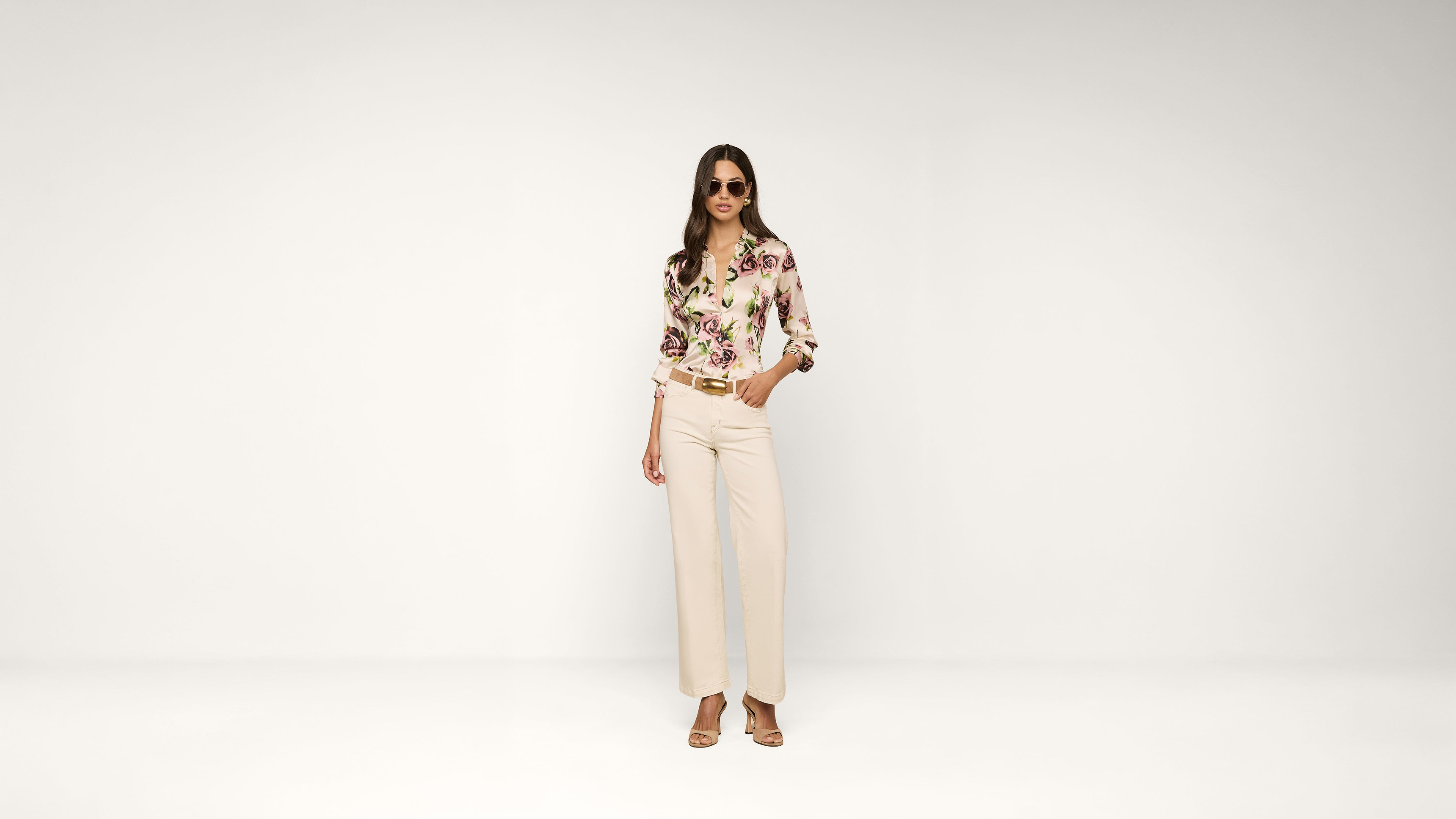 A woman wearing sunglasses, a floral blouse, cream wide-leg pants, and heeled sandals stands confidently against a plain white background.