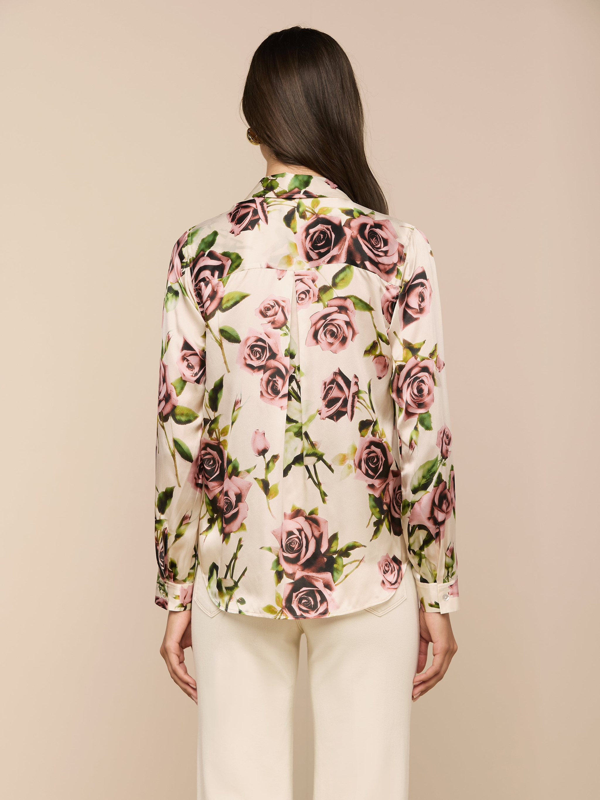 Una donna con lunghi capelli castani, vista da dietro, indossa la blusa di seta LAGENCE Tyler - un bottone color crema con una stampa di rose color malva - e pantaloni bianchi su uno sfondo beige.