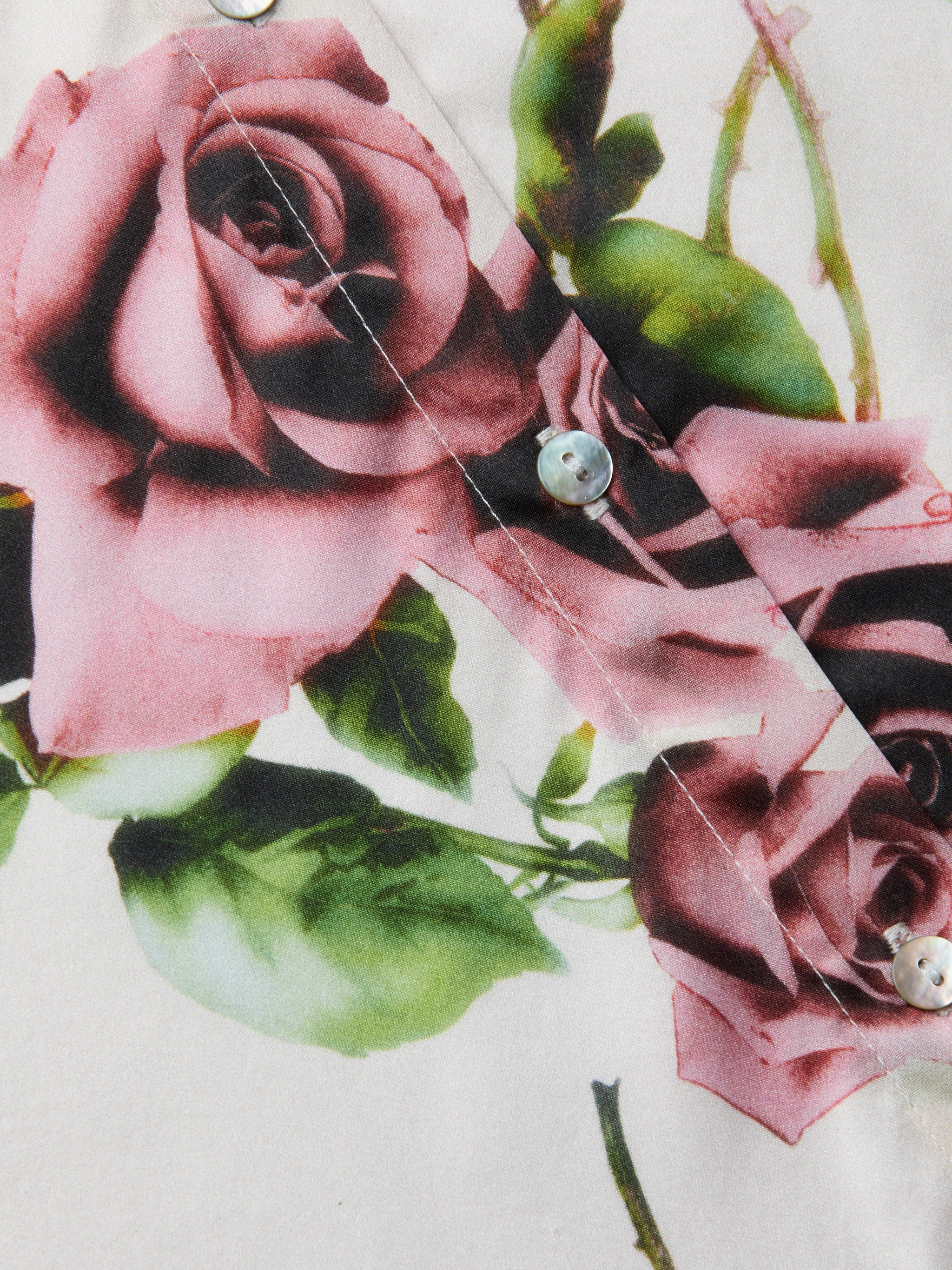 Primo piano del tessuto stampato a rose color malva con foglie verdi, con una cucitura ornata da bottoni iridescenti: dettagli classici della blusa in seta Tyler di LAGENCE.