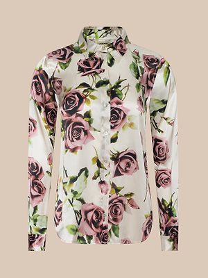 The Tyler Silk Blouse by LAGENCE is a long-sleeved, collared button-down in white silk with a mauve rose and green leaf print, shown against a beige background.