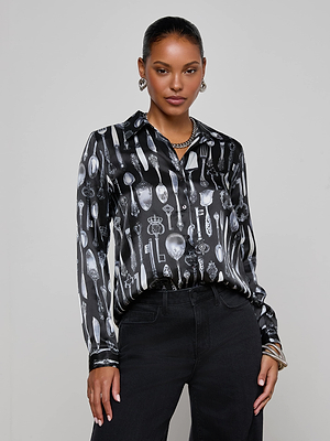 A woman poses confidently against a plain background wearing the L'AGENCE Tyler Silk Blouse, featuring a black fabric with a white silverware print, paired with black pants. She accessorizes with hoop earrings, a necklace, bracelets, and has slicked-back hair.