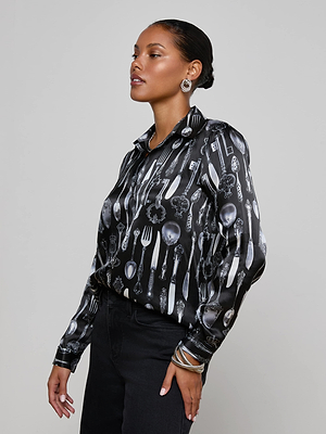 A woman in the L'AGENCE Tyler Silk Blouse, featuring a silverware print of spoons, forks, and knives, stands against a plain background. She wears statement earrings and bangles with slicked-back hair, looking confidently to the side.