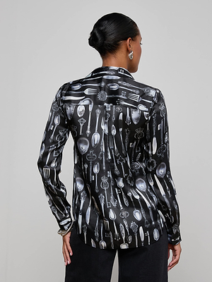 A person faces away, wearing the L'AGENCE Tyler Silk Blouse in a black silverware print—forks, spoons, and knives—with dark pants against a plain light gray background.