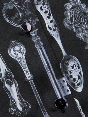 Close-up of the L'AGENCE Tyler Silk Blouse in black, showcasing a white vintage silverware print—ornate spoons and forks—and two black buttons on its luxurious fabric.