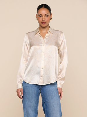 A woman stands facing forward against a plain beige background, wearing the Tyler Silk Blouse—a light, button-down top with a subtle paisley print—and blue jeans.