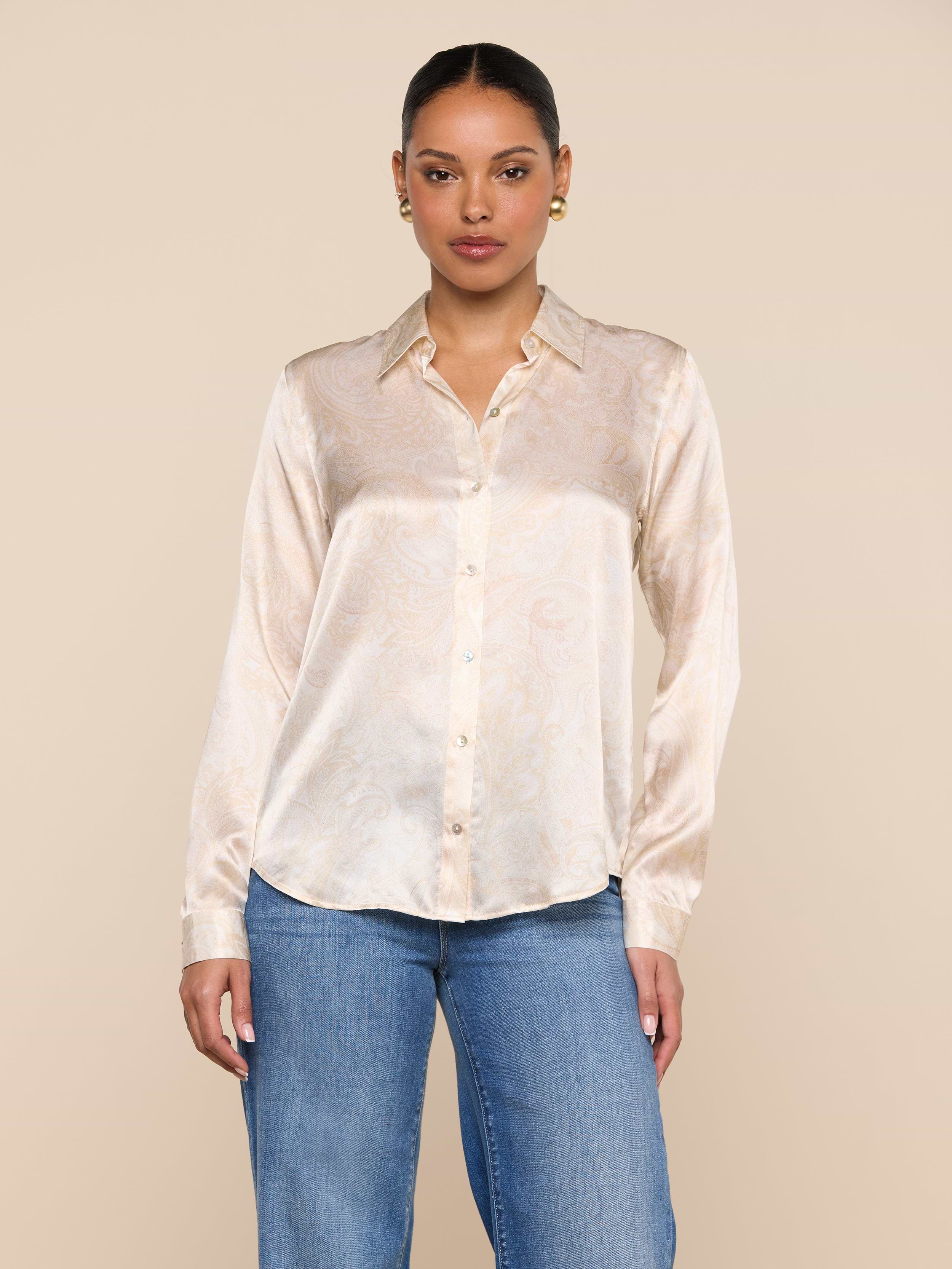 A woman wearing the Tyler Silk Blouse in light beige with a subtle paisley print, paired with blue jeans, stands against a plain off-white background.