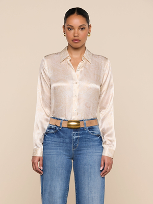 A woman faces forward wearing the Tyler Silk Blouse in light beige with a subtle paisley print, paired with blue jeans, a wide beige belt featuring a large gold buckle, and gold earrings. Her hair is pulled back and her expression is neutral.